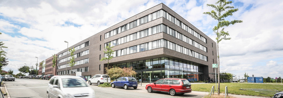 Informatik-ITMC ITMC: Ein großes, braun-rot verklinkertes Gebäude mit Flachdach aus der Froschperspektive fotografiert. Davor eine Straße, auf der ein Auto fährt. Andere Autos stehen am Straßenrand hintereinander geparkt.